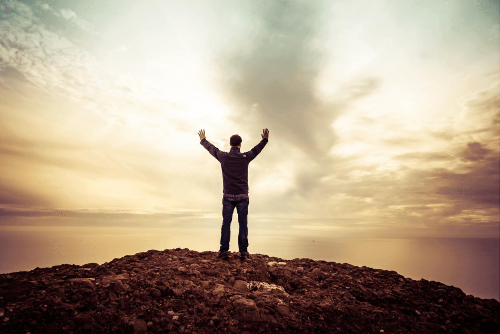 Person standing on rocky hill with arms raised, overlooking a cloudy sunrise.